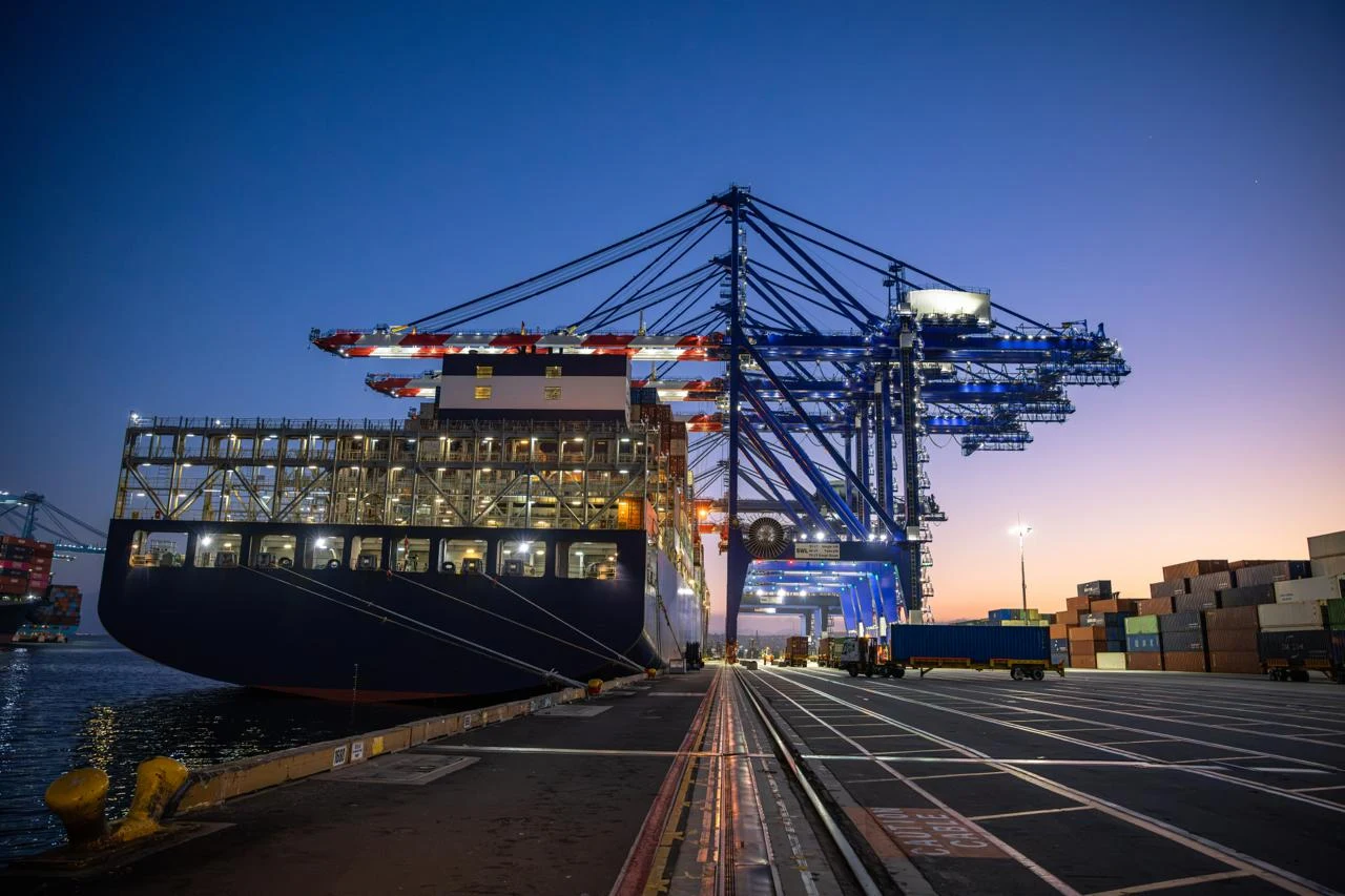 A large container ship berthed under gantry cranes at a US port at night, lit against a deep-blue dusk sky