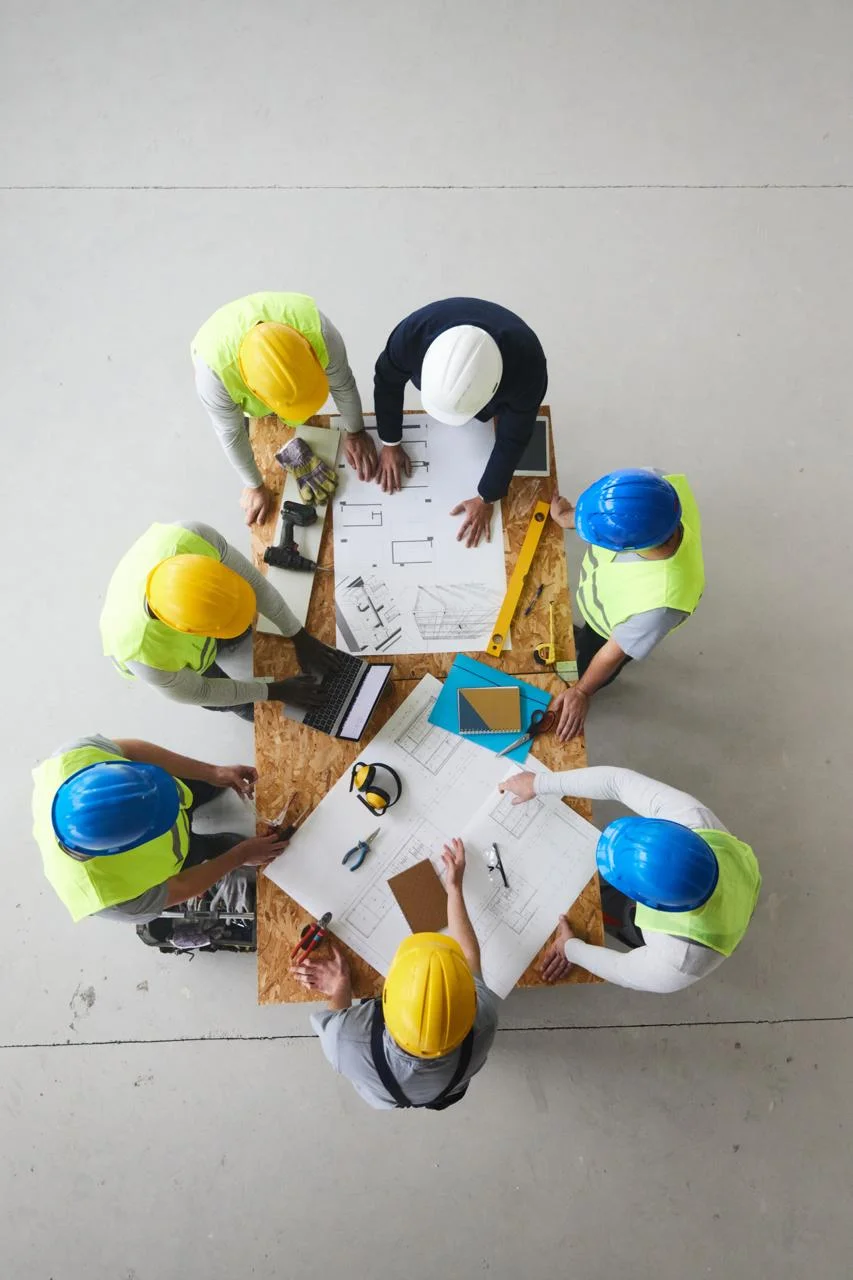 Top-down view of a construction team gathered around a table reviewing blueprints and a laptop.