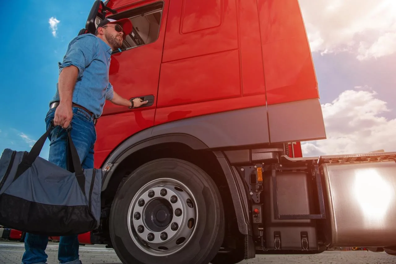 Truck driver with a duffel bag boarding the cab of a red semi at sunset — the end-of-shift moment Globeo-managed lodging fits into